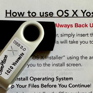 May include: A silver USB flash drive with a black USB connector. The drive has a white label that says "The ComPlace 10.10 Yosemite USB 3.0". The drive is sitting on a white piece of paper with instructions on how to use OS X Yosemite.