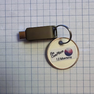May include: A gray USB-C flash drive attached to a keyring with a round tag. The tag has the text "The ComPlace 12 Monterey" and a graphic of a circle with red, white, and blue colors. The items are on a grid background.