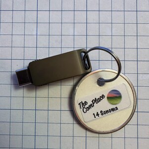 May include: A silver-colored key ring with a round tag and a dark gray USB-C flash drive. The tag has a sticker with the text "The ComPlace 14 Sonoma" and a landscape graphic. The flash drive is connected to the key ring.