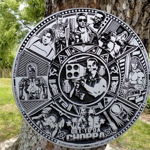 May include: A circular metal plaque with a black and white design featuring various characters and symbols. The plaque has a raised border and a central image of a man holding a weapon. The text "GET TO DA CHOPPA" is printed at the bottom of the plaque.