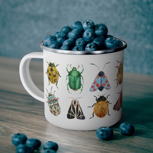 May include: White enamel mug with a colorful insect design. The mug features a variety of insects, including beetles, moths, and butterflies. The mug is filled with blueberries.