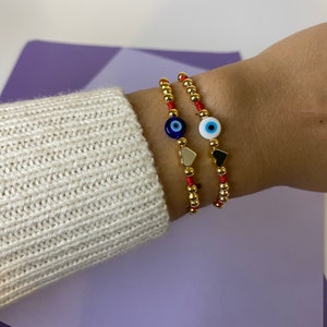 May include: Two red string bracelets with gold beads and evil eye charms. One charm is blue and white, the other is blue and black.