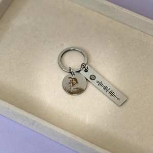 May include: Silver keychain with a round charm bearing a couple's photo and a rectangular tag with a Spotify code. The keychain is presented in a light-coloured tray.