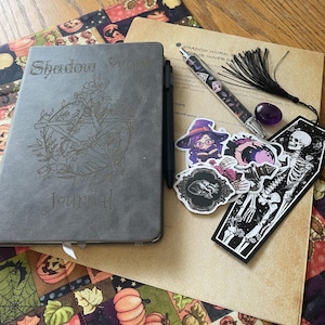 May include: A gray journal with the words "Shadow Work Journal" and a floral design, a pen, stickers, and a decorative tag with a skeleton design. The items are arranged on a patterned fabric with pumpkins and sunflowers.