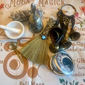 May include: An assortment of spiritual items including a white mortar and pestle, a sage bundle, a decorative bell, a small broom, and a pocket watch with a celestial design. The items are arranged on a cloth with the words "Moon Magick".