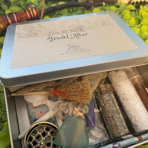 May include: A silver metal box with the text "Sea Witch Travel Altar" and the words "The Wicked Grimoire" on the lid. The box is open and contains a variety of items, including a small broom, seashells, a starfish, a glass bottle, and a small metal container.