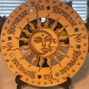 May include: A wooden planchette board with a sun and moon design. The board has the words "Yes", "No", and "Rephrase" along the perimeter, along with the alphabet and numbers. The board is displayed on a black stand.