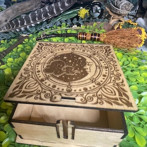 May include: A wooden box with an open lid, featuring intricate laser-cut celestial designs. The box is light brown with a detailed pattern of stars, moons, and swirling clouds. The box is sitting on a bed of green leaves.