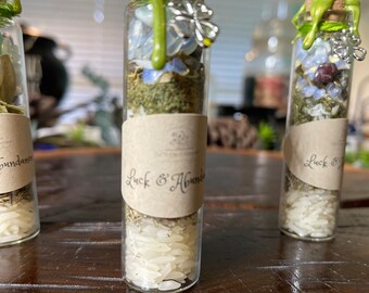 May include: Three small glass bottles filled with herbs, crystals, and rice. The bottles are sealed with green wax and have a small silver butterfly charm attached. The labels on the bottles read "Abundance", "Luck & Abundance", and "Luck & Good Fortune".