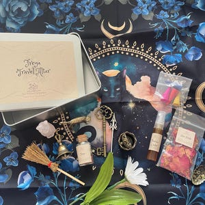 May include: A silver tin box with the text "Freya Travel Altar" on the lid. The box is open and contains a variety of items, including a small broom, a bell, a bottle of liquid, a key, a small cat figurine, a crystal, and a small bag of dried rose petals. The items are arranged on a black fabric with a floral pattern and a moon and stars design.