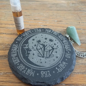 May include: A black slate coaster with a crescent moon and crystal design, surrounded by text. A green crystal pendulum and a small bottle of liquid are also present. The coaster is approximately 4 inches in diameter.