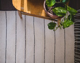 May include: A white rug with black stripes and fringe. The rug is on a wooden floor and a potted plant is in the upper right corner of the image.