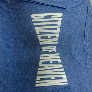 May include: Blue t-shirt with the text "CITIZEN OF HEAVEN" in a white, hourglass-shaped design. The shirt is made of a soft, textured fabric. The text is centered vertically.