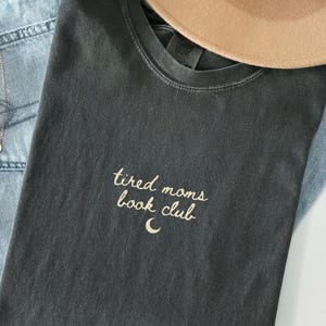 May include: Dark gray t-shirt with the text "tired moms book club" and a crescent moon graphic. The shirt is paired with light blue jeans, a tan hat, and a silver necklace. The shirt is made of a soft, comfortable material.