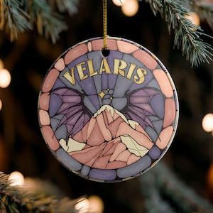 Könnte beinhalten: Runder Weihnachtsschmuck mit dem Wort "VELARIS" in goldfarbenen Buchstaben. Der Schmuck zeigt eine Berglandschaft in Lila und Rosa, umrahmt von einem Buntglas-Stil-Rand. Er hängt an einer goldenen Schnur.