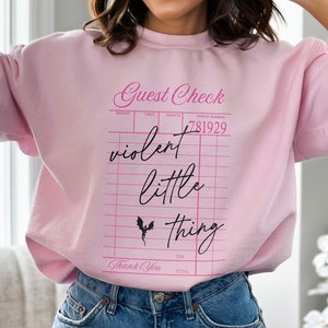 May include: Pink sweatshirt with a graphic of a guest check that says "violent little thing" in pink script with a black bat graphic.