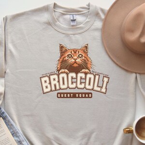 Pu&ograve; includere: Una felpa beige chiaro con una grafica marrone di un gatto che sporge sopra il testo "BROCCOLI QUEST SQUAD". Il testo "no one wants it" &egrave; a sinistra della grafica e il testo "but it's good for you" &egrave; a destra della grafica.