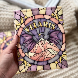 May include: A book sleeve with a stained glass design featuring a mountain range, crescent moons, and stars. The text "VELARIS" is written in gold lettering.