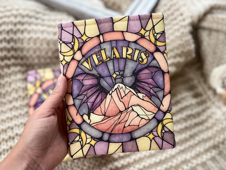 May include: A book sleeve with a stained glass design featuring a mountain range, crescent moons, and stars. The text "VELARIS" is written in gold lettering.