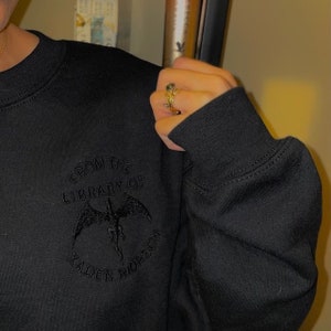 K&ouml;nnte beinhalten: Schwarzer Pullover mit einem schwarzen gestickten Design, das "From the Library of Kaden Riderson" mit einer schwarzen Drachengrafik sagt.