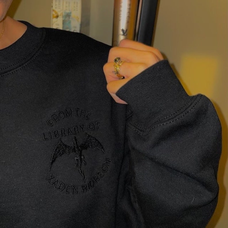 K&ouml;nnte beinhalten: Schwarzer Pullover mit einem schwarzen gestickten Design, das "From the Library of Kaden Riderson" mit einer schwarzen Drachengrafik sagt.