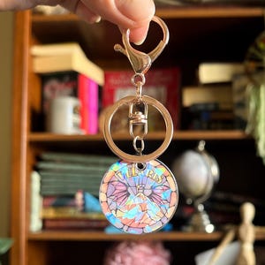 Op de afbeelding: Een gouden sleutelhanger met een ronde, iriserende hanger met een berglandschap en de tekst "Tel Aviv".