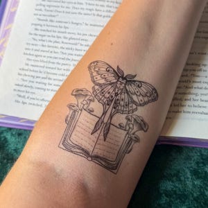 May include: A black ink temporary tattoo on skin featuring a detailed moth with patterned wings positioned above an open book. Several small mushrooms grow from the pages and spine of the book, rendered in a fine line art style with shading.