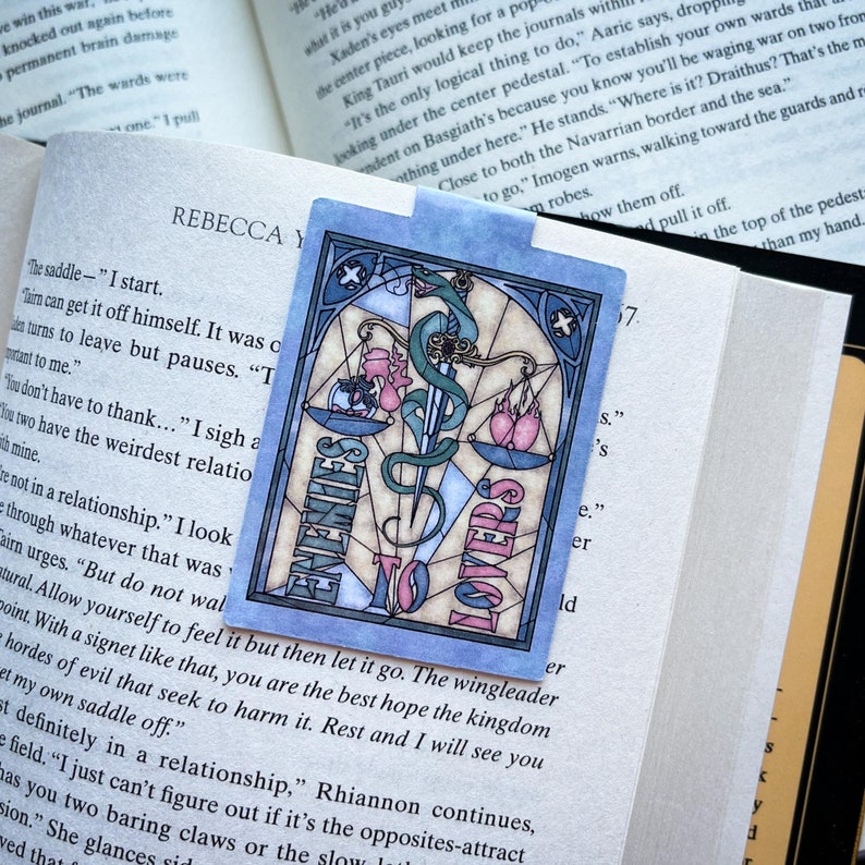 K&ouml;nnte beinhalten: Ein blaues und rosafarbenes Lesezeichen mit einem Buntglasdesign einer Waage mit einem Schwert und den Worten "Enemies to Lovers".