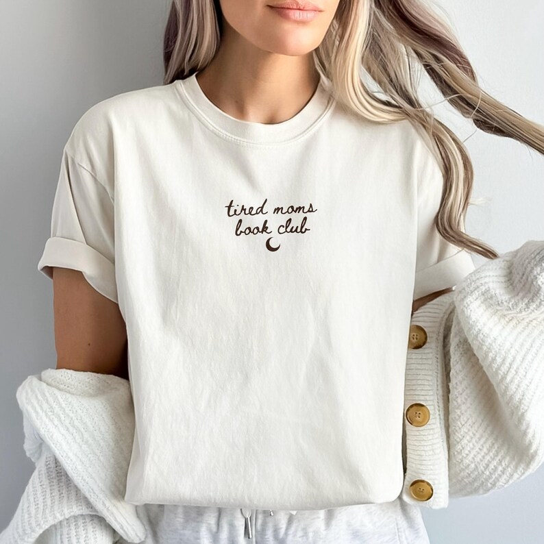 May include: Cream-colored t-shirt with the text "tired moms book club" in a dark brown script, accompanied by a crescent moon graphic. The shirt has rolled-up sleeves and a relaxed fit, suggesting comfort and casual wear.