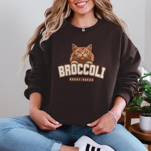 Pu&ograve; includere: Una felpa nera con un gatto marrone che spunta dalla parte superiore. La felpa ha la scritta "Broccoli Quest Squad" stampata sul davanti.