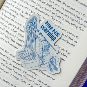 May include: A die-cut sticker featuring a blue and white illustration of a medieval scene. A woman in a long gown holds a sword over a kneeling figure. The sticker has the text "bring back yearning".
