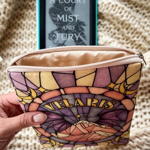 May include: A beige zippered pouch with a stained glass design featuring a purple, pink, and yellow color scheme. The pouch has the words "Velaris" printed in gold lettering.