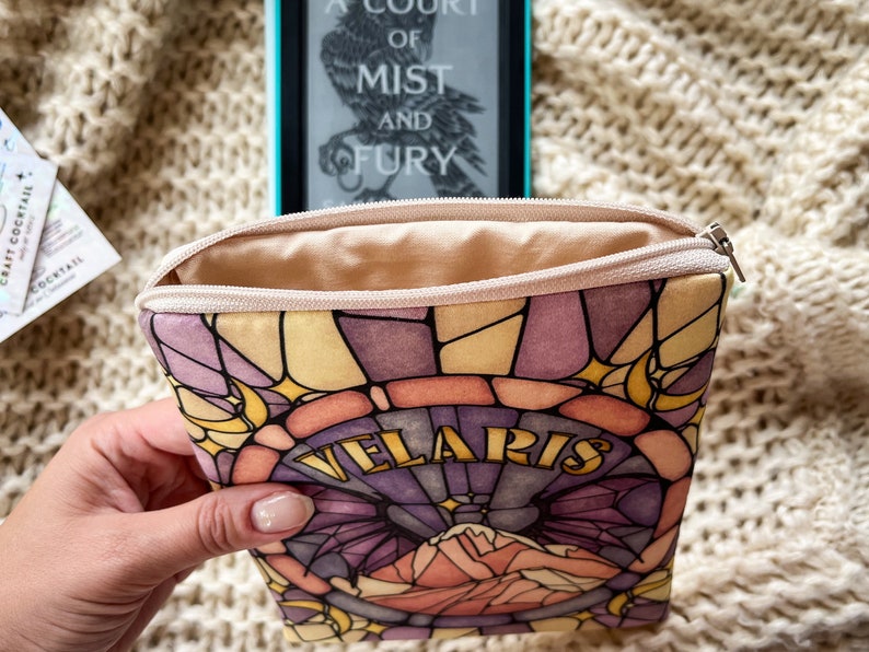 May include: A beige zippered pouch with a stained glass design featuring a purple, pink, and yellow color scheme. The pouch has the words "Velaris" printed in gold lettering.