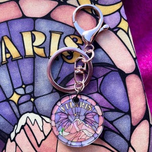 Op de afbeelding: Een sleutelhanger van zilver en ros&eacute;goud met een ronde glazen hanger met een glas-in-loodontwerp van een bergketen met het woord "Velaris" in gouden letters.