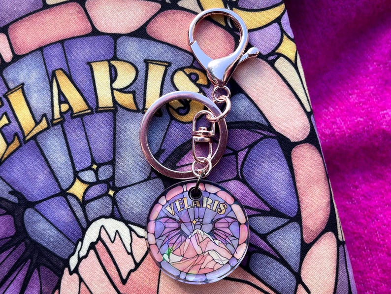 Op de afbeelding: Een sleutelhanger van zilver en ros&eacute;goud met een ronde glazen hanger met een glas-in-loodontwerp van een bergketen met het woord "Velaris" in gouden letters.