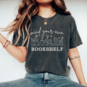 May include: Dark gray t-shirt with the text "mind your own bookshelf" in white script. The design includes a bookshelf illustration with flowers and books. The shirt is a casual, comfortable style.