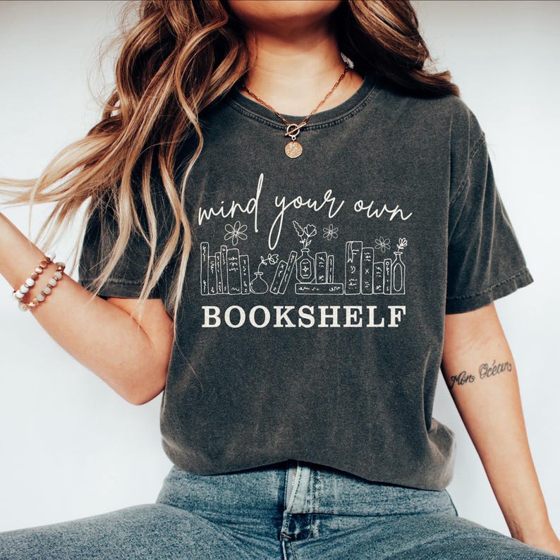 May include: Dark gray t-shirt with the text "mind your own bookshelf" in white script. The design includes a bookshelf illustration with flowers and books. The shirt is a casual, comfortable style.