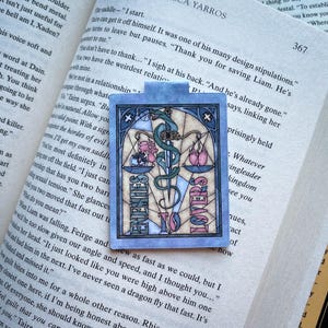 K&ouml;nnte beinhalten: Ein blaues und rosafarbenes Lesezeichen mit einem Buntglasdesign. Das Lesezeichen zeigt eine Waage mit einem Schwert und einer Schlange. Die Worte "Enemies to Lovers" sind auf dem Lesezeichen geschrieben.