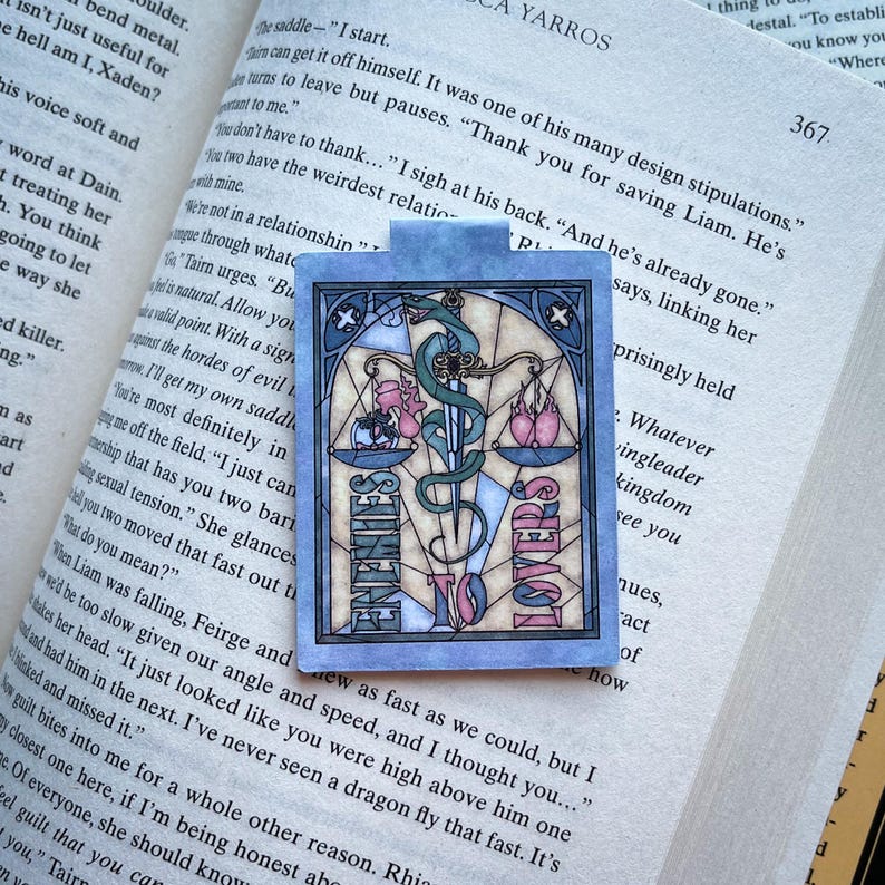 K&ouml;nnte beinhalten: Ein blaues und rosafarbenes Lesezeichen mit einem Buntglasdesign. Das Lesezeichen zeigt eine Waage mit einem Schwert und einer Schlange. Die Worte "Enemies to Lovers" sind auf dem Lesezeichen geschrieben.