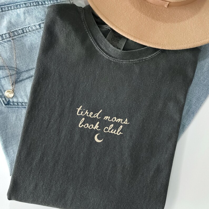 May include: Dark gray t-shirt with the text "tired moms book club" and a crescent moon graphic. The shirt is paired with light blue jeans, a tan hat, and a silver necklace. The shirt is made of a soft, comfortable material.