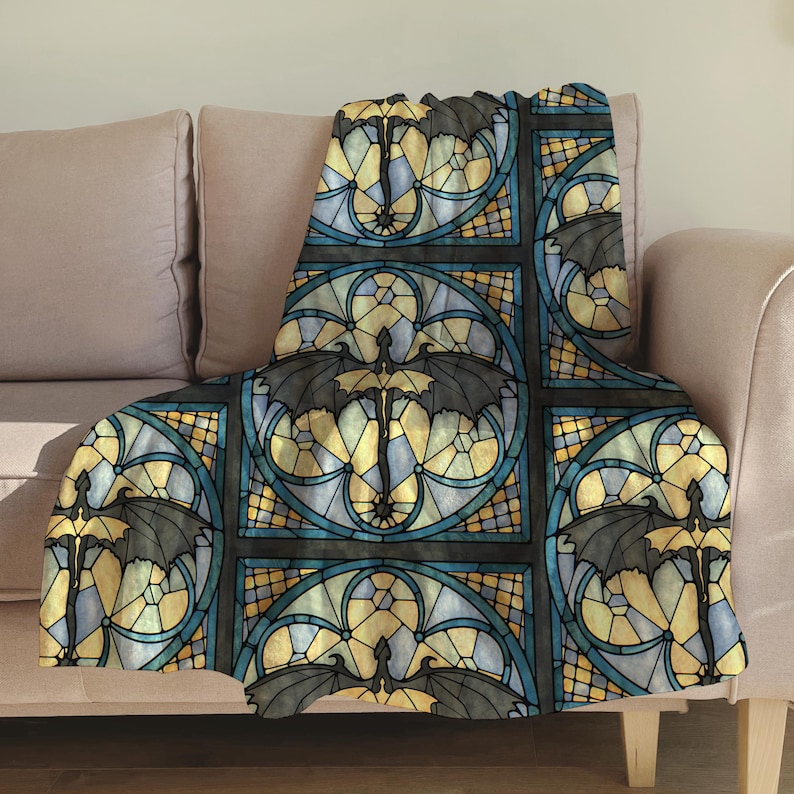 May include: A decorative throw blanket featuring a stained-glass design with a repeating pattern of bats. The design incorporates black, yellow, and blue colors, creating a gothic aesthetic. The blanket is draped over a light pink sofa.