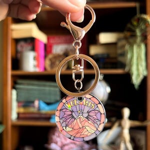 Op de afbeelding: Een gouden sleutelhanger met een ronde, kleurrijke, glas-in-lood stijl hanger met een bergketen en het woord "Velaris".