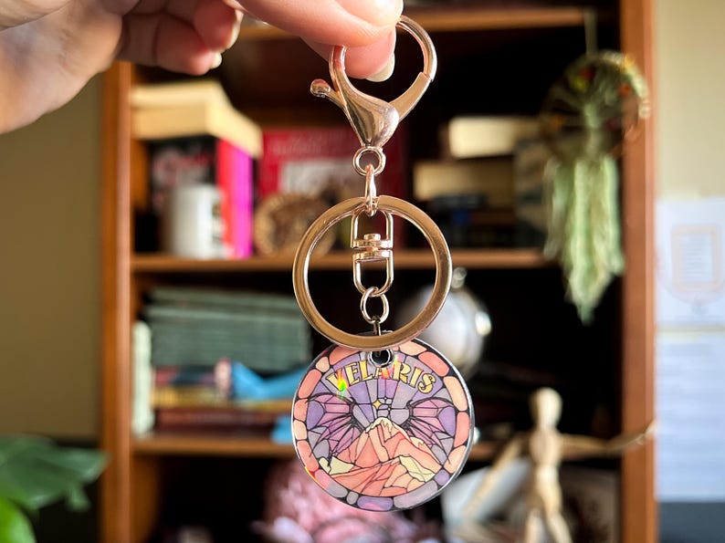 Op de afbeelding: Een gouden sleutelhanger met een ronde, kleurrijke, glas-in-lood stijl hanger met een bergketen en het woord "Velaris".