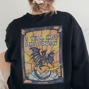 Pu&ograve; includere: Felpa nera con un design a vetri colorati che presenta un drago e il testo "I WILL NOT DIE TODAY".