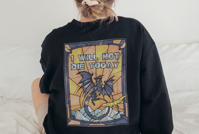 Pu&ograve; includere: Felpa nera con un design a vetri colorati che presenta un drago e il testo "I WILL NOT DIE TODAY".