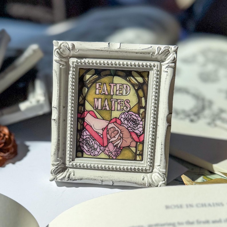 May include: A small, rectangular framed art piece with a distressed white finish. The artwork features the words "FATED MATES" above a design of hands, roses, and ribbons in pink and yellow tones. The frame has ornate detailing.