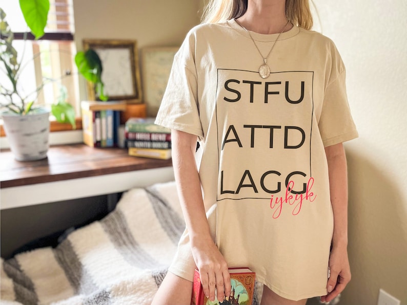 May include: A beige t-shirt with a black square graphic that says "STFU ATTD LAGG" in black text and "iykyk" in pink text.