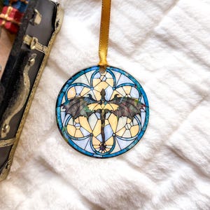 Op de afbeelding: Ronde ornament met een glas-in-lood ontwerp met een draak met uitgespreide vleugels. Het heeft een gouden lint om op te hangen. Het ontwerp gebruikt blauwe, gele en zwarte kleuren, wat een opvallend visueel effect cre&euml;ert. Waarschijnlijk voor feestelijke of decoratieve doeleinden.