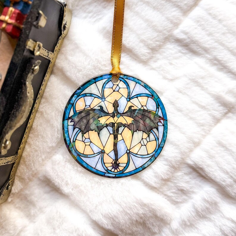 Op de afbeelding: Ronde ornament met een glas-in-lood ontwerp met een draak met uitgespreide vleugels. Het heeft een gouden lint om op te hangen. Het ontwerp gebruikt blauwe, gele en zwarte kleuren, wat een opvallend visueel effect cre&euml;ert. Waarschijnlijk voor feestelijke of decoratieve doeleinden.