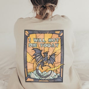 Pu&ograve; includere: Una felpa beige con un designo a vetri colorati che presenta un drago blu e il testo "I WILL NOT DIE TODAY".